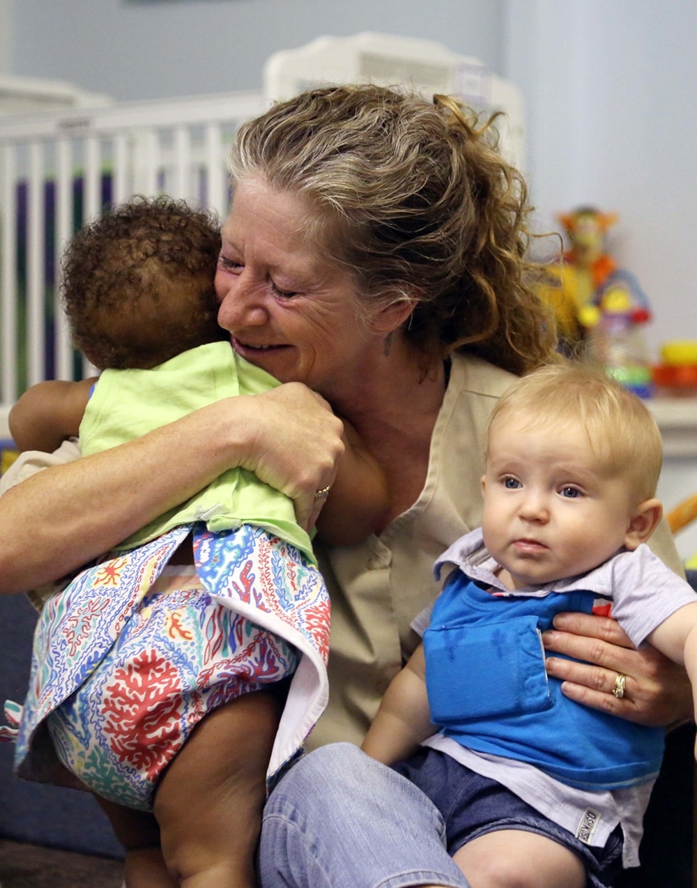 A smiling woman hugs a baby with curly hair while holding another baby with blond hair on her lap in a colorful, indoor setting.