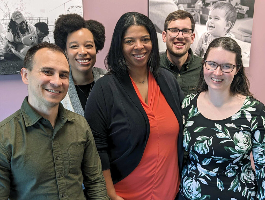 Five adults stand together smiling in front of a pink wall with black-and-white photos of children. They appear friendly and relaxed, dressed in casual professional clothing.