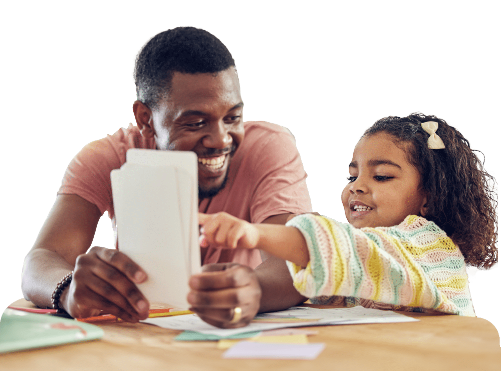 A smiling man holds up flashcards while a young girl with curly hair excitedly points at one. They sit at a table together, appearing to enjoy a learning activity.