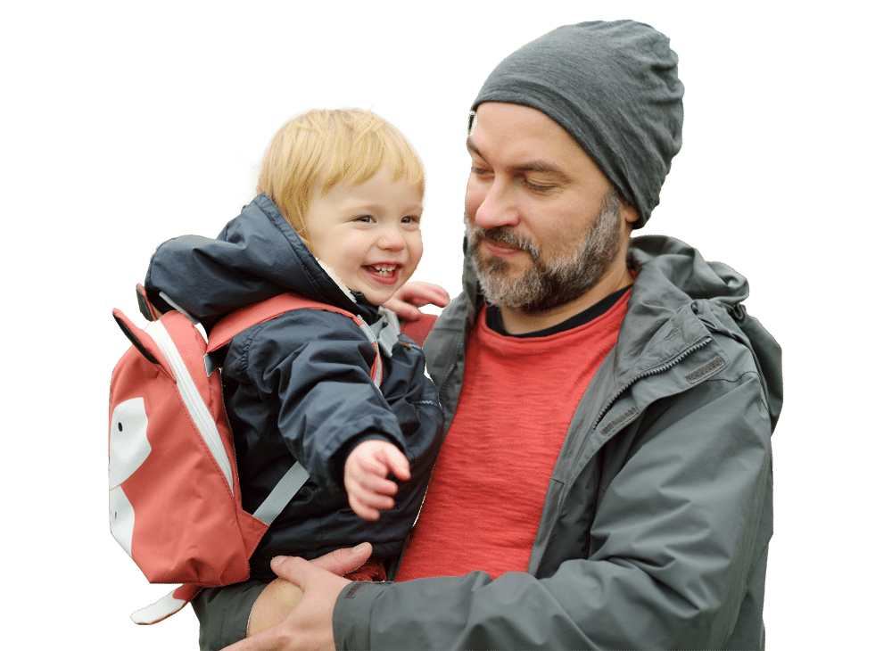 A man in a gray beanie and jacket holds a smiling toddler wearing a raincoat and a pink backpack. The man looks at the child fondly against a plain, light background.