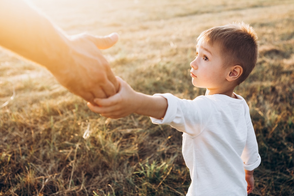 A young boy in a white shirt holds an adult’s hand while looking up, standing outdoors in a sunlit, grassy field. The scene conveys warmth, trust, and connection.