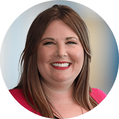 A woman with long, straight brown hair smiles at the camera. She is wearing a bright pink top and is pictured against a softly blurred background.