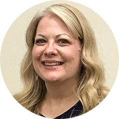 Smiling woman with long, wavy blonde hair, wearing a dark top, photographed against a plain, light-colored background.