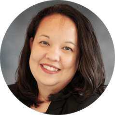 A woman with long dark hair and a friendly smile, wearing a black top, poses against a neutral gray background.