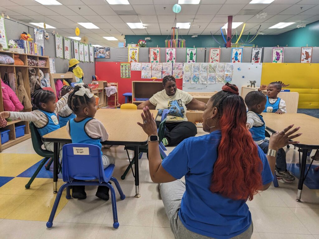 Two adults lead a group activity with six young children in a colorful, language rich environment. The kids wear blue vests as they engage enthusiastically, surrounded by art projects that decorate the walls.