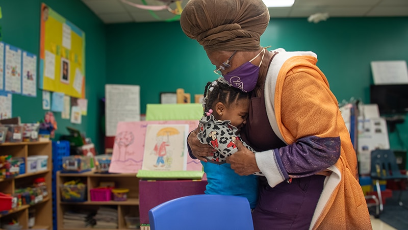 A woman wearing a headwrap and mask lovingly hugs a young child in a colorful classroom filled with artwork, books, and educational materials.