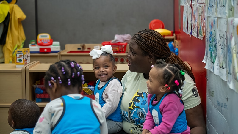 A caregiver sits on the floor in a playroom, surrounded by young children wearing blue aprons. One smiling girl with white bows in her hair is sitting on the caregiver’s lap. Colorful drawings hang on the wall.
