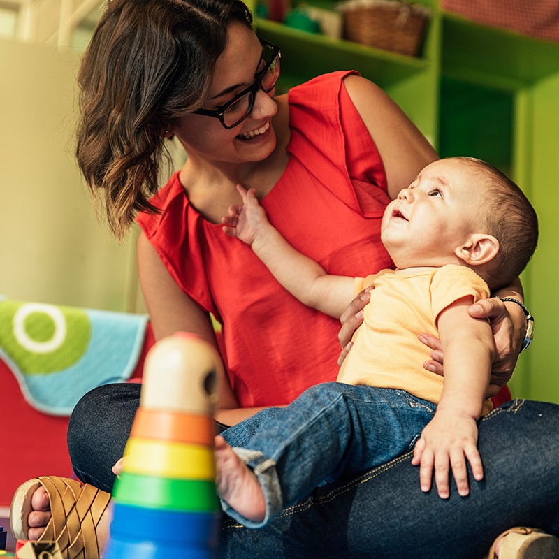 A smiling woman in glasses holds a baby on her lap in a colorful playroom. The baby reaches towards her, and a wooden rainbow stacking toy is in the foreground.