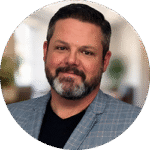 A man with short dark hair and a trimmed beard, wearing a gray plaid blazer over a black shirt, smiles slightly in a softly blurred indoor setting.