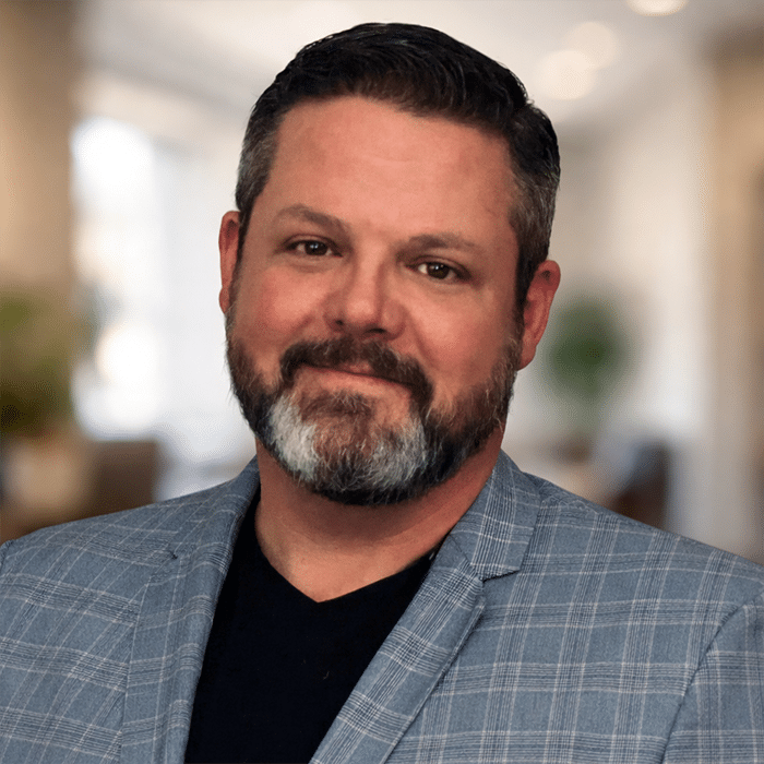 A man with short dark hair and a beard with gray highlights, wearing a gray plaid blazer over a black shirt, smiles in a softly lit, blurred indoor setting.