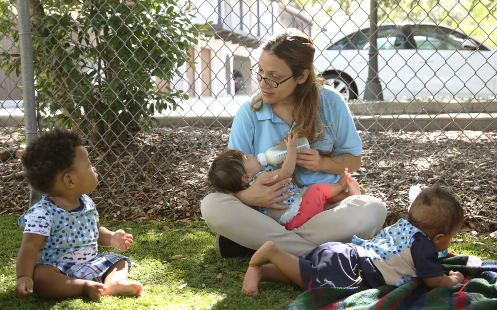 A woman sits on grass feeding a baby with a bottle while two other small children sit nearby, one on a blanket, engaging in outdoor play and talking together in an outdoor fenced area.