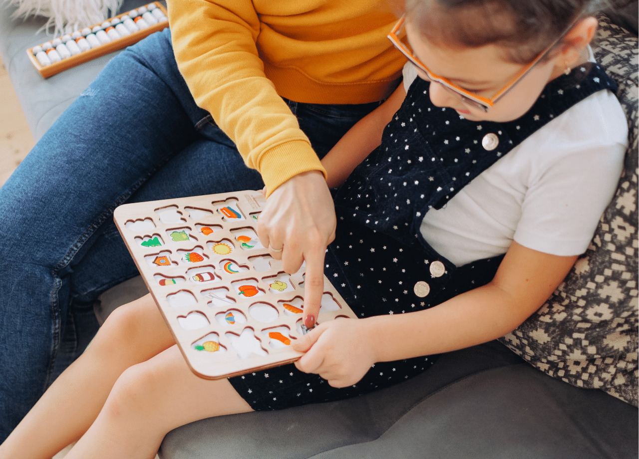 An adult and a child sit on a couch, using a wooden educational puzzle with colorful pieces. The adult points to the puzzle, while the child holds and examines it closely.