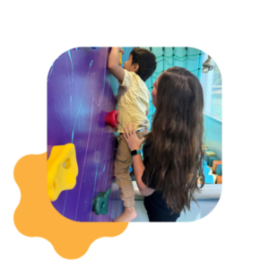 A woman supports a young child as he climbs a colorful indoor rock wall. Bright holds and playful room decor are visible in the background.