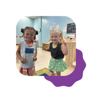 Two young children smiling and posing together in a brightly lit classroom, both wearing colorful outfits. A purple shape decorates the bottom right corner of the image.