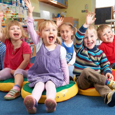 Six young children sit on colorful cushions in a classroom, smiling and raising their hands enthusiastically. The background shows toys, a desk, and classroom decorations. The kids appear happy and engaged.