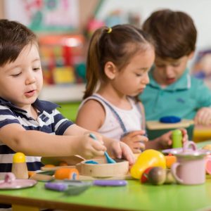 Three young children sit at a table playing with colorful wooden toy food and kitchen utensils in a bright classroom or daycare setting.