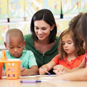 A teacher sits at a table with young children, helping them with a coloring activity in a classroom setting. The kids are focused on their drawings, using crayons and paper.