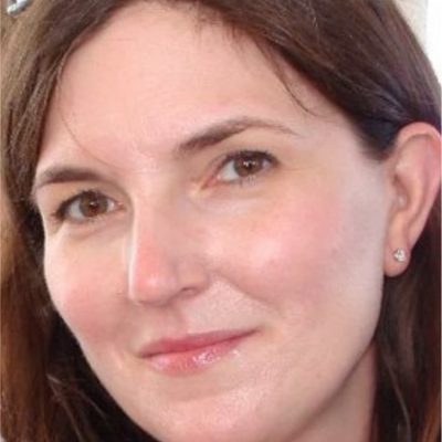 A close-up photo of a woman with light skin, brown hair, and brown eyes, looking at the camera with a slight smile. She is wearing a small stud earring and has a natural, minimal makeup look.