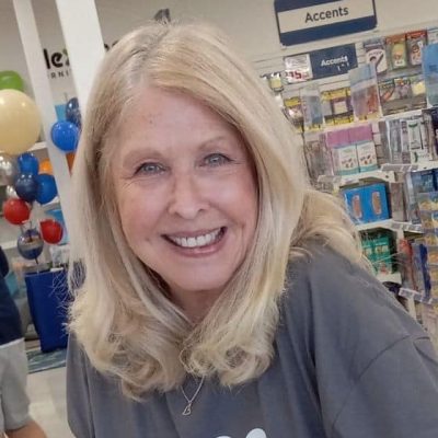 A smiling older woman with long blonde hair stands indoors near shelves of merchandise and a sign labeled "Accents." Balloons and store displays are visible in the background.