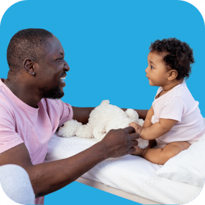 A smiling man in a pink shirt plays with a baby sitting on a bed, holding a white stuffed animal. The baby faces him, and both appear happy, against a solid blue background.