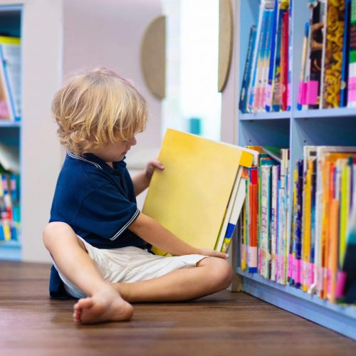 A young child with blonde hair sits barefoot on the floor by a bookshelf, looking at a large yellow book in a brightly lit library or classroom.