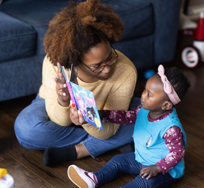 2022.03.R5.0251_3000px-scaled A mother sits cross-legged on the floor, holding up a colorful book and smiling at a young toddler girl beside her. The girl, wearing a headband, looks at the book and reaches toward it. They are in a cozy living room.