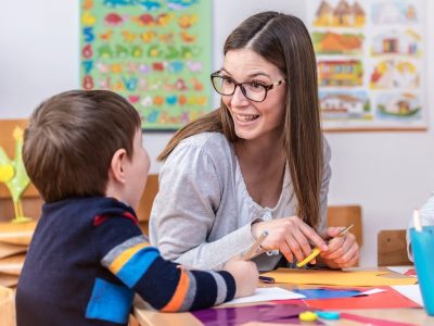 A woman with glasses smiles and talks to a young boy at a classroom table. They are doing arts and crafts with colorful paper. Educational posters are visible on the wall in the background.