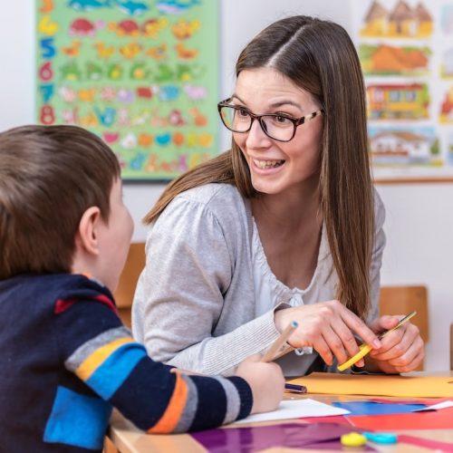 A woman with glasses smiles and talks to a young boy at a classroom table. They are doing arts and crafts with colorful paper. Educational posters are visible on the wall in the background.