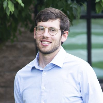 A man with short brown hair, glasses, and a beard smiles at the camera. He is wearing a light blue collared shirt and standing outdoors near greenery and a building with large windows.