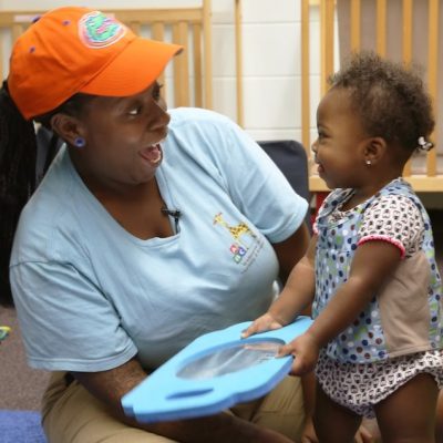 An adult and a toddler smile at each other while playing with a blue toy in a nursery, with cribs and toys visible in the background.