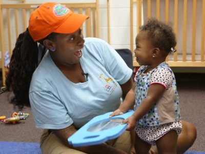 An adult and a toddler smile at each other while playing with a blue toy in a nursery, with cribs and toys visible in the background.