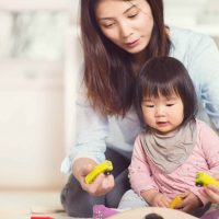 An adult woman and a young girl sit on the floor, playing together with yellow toy cars. The woman gently guides the child, who is focused on the toys. The background shows a bright, softly lit room.