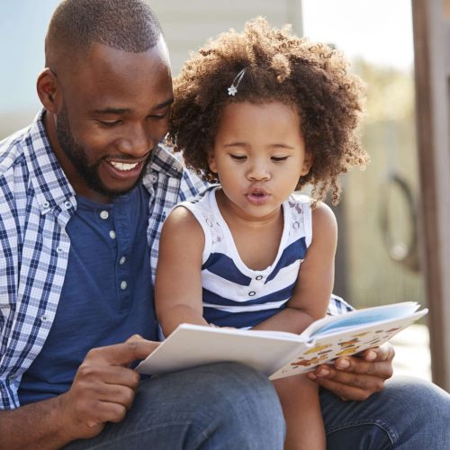 A man and a young girl sit outside, smiling as they read a book together. The man points at the pages while the girl, wearing a striped dress, looks at the book, engaged and focused.