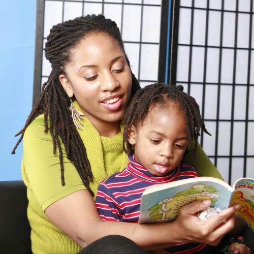 A woman with long braids smiles while reading a colorful children’s book to a young child sitting on her lap. They are sitting against a black and white folding screen.
