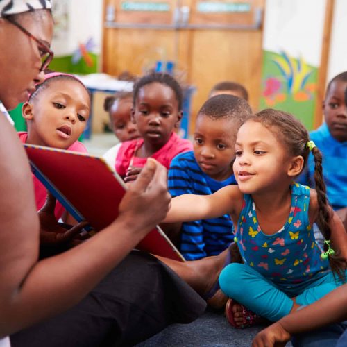 A teacher sits on the floor reading a book to a group of young children who are gathered closely around her, listening attentively and looking at the book with interest.