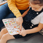 An adult and a child sit on a couch, using a wooden educational puzzle with colorful pieces. The adult points to the puzzle, while the child holds and examines it closely.