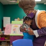 A woman wearing a headwrap and mask lovingly hugs a young child in a colorful classroom filled with artwork, books, and educational materials.