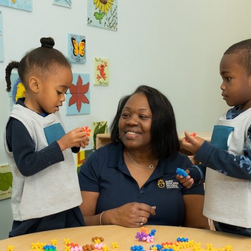 A teacher sits at a table with two young children, all smiling and holding colorful plastic building toys. Behind them are flower-themed art pieces and bookshelves filled with books.