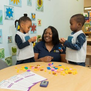 A teacher sits at a round table with two young children, who are playing with colorful star-shaped counters. Educational materials and flower artwork decorate the classroom walls in the background.