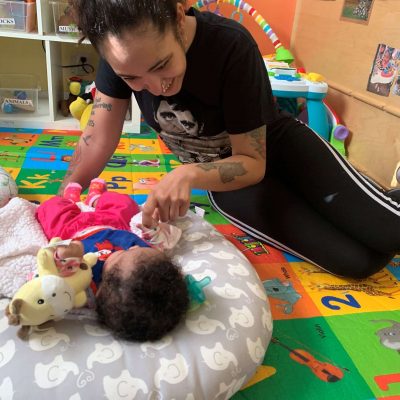 KB-infant-with-her-mom A woman smiles and interacts with a baby lying on a cushion in a colorful playroom with alphabet and number mats. The baby reaches for a soft toy while the woman leans in close and looks happy.
