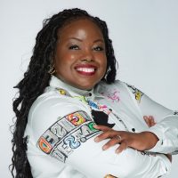 A smiling woman with long braided hair poses confidently with arms crossed, wearing a white shirt decorated with colorful patches and designs, against a plain white background.