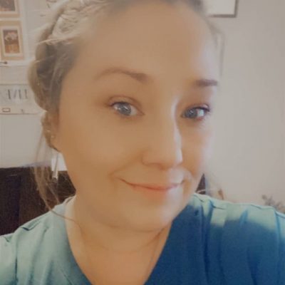 A woman with braided hair and light makeup smiles at the camera, wearing a teal top and necklace. The background shows framed pictures and documents on a light-colored wall.