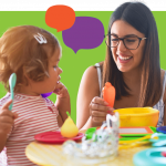 A young woman wearing glasses smiles while sitting at a table with a toddler holding a spoon. Colorful dishes and toy food are on the table, with speech bubble graphics in the background.