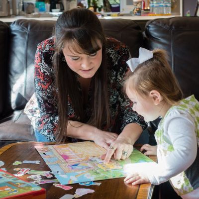 A woman and a young girl work on a colorful puzzle together at a wooden table in a cozy living room with brown sofas and cityscape pillows. Sunlight streams in, highlighting the puzzle pieces.