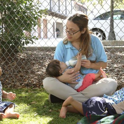 A woman sits on grass outdoors, bottle-feeding a baby in her lap while two other young children sit nearby, all surrounded by a chain-link fence and trees.