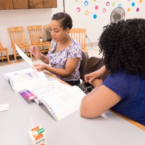 Two women sit at a table in a classroom, looking at and discussing documents or books. One woman holds a paper, while the other listens. Chairs and colorful wall decorations are visible in the background.