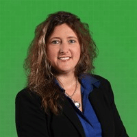 A woman with wavy brown hair wearing a dark blazer over a blue shirt and necklace, smiling in front of a bright green background.