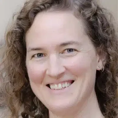 A woman with curly brown hair and blue eyes smiles at the camera. She is wearing a small earring and appears in a softly lit indoor setting.