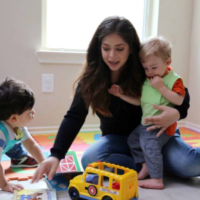 Mom_With_Boys_LENA_Day_1 A woman sits on the floor with two young children, playing with toys including a yellow school bus and some books, on a colorful mat in a bright room with a window.
