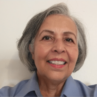 An older woman with short gray hair smiles at the camera. She is wearing a light blue collared shirt and is standing in front of a plain white background.